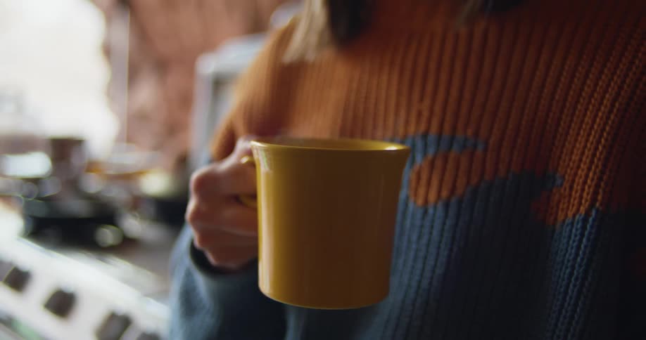 Woman wearing sweater stands in kitchen holding steaming coffee mug