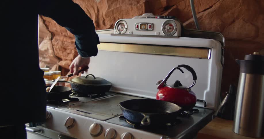A man scrapes a cast iron pan with a spatula in a kitchen with a rock wall