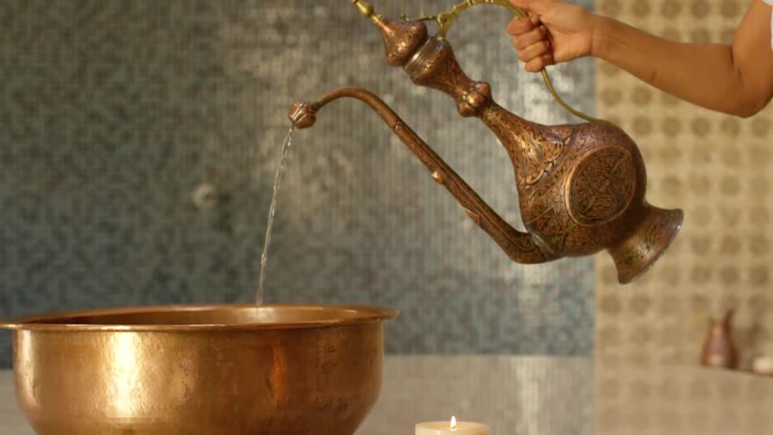 A person pouring a pitcher of water in a basin on a table with a candle