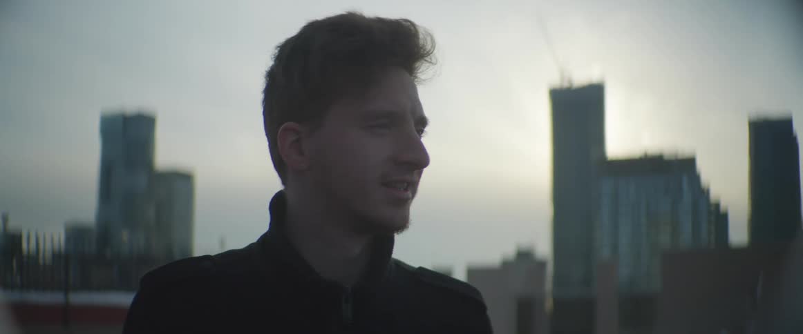 A man on a rooftop with a city skyline buildings the sky and clouds in the background