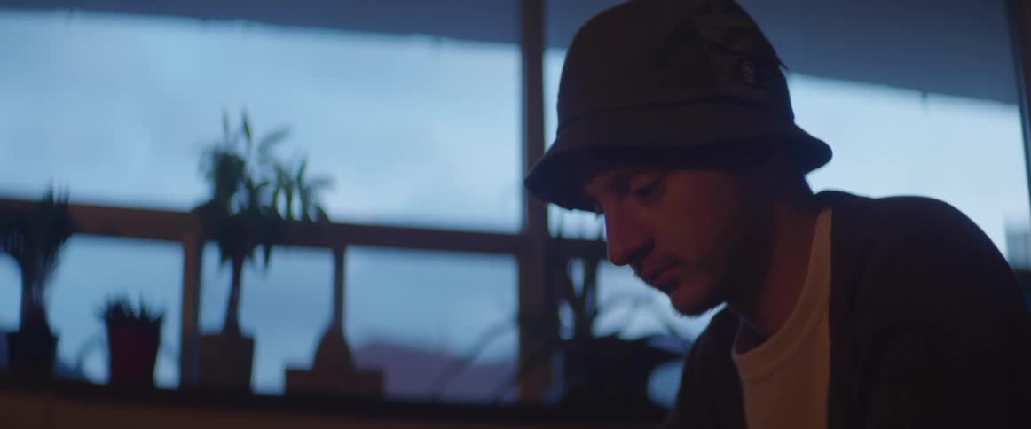 Man wearing hat sits looking down next to window lined with houseplants