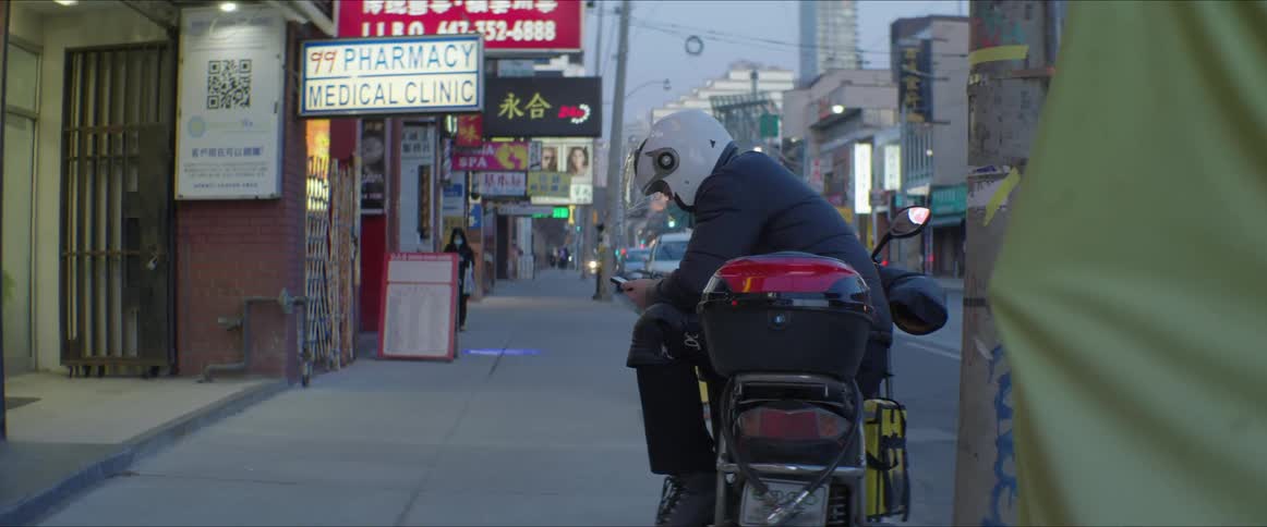 A person with a smart phone and a helmet sitting on a motorcycle with buildings the sky and clouds in the background