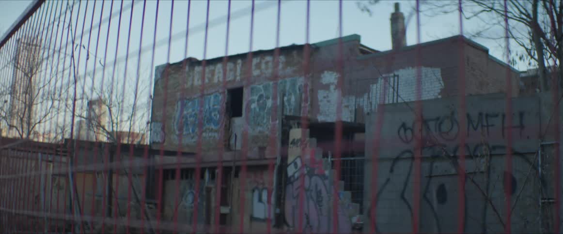 A fence with buildings with graffiti the sky and clouds in the background