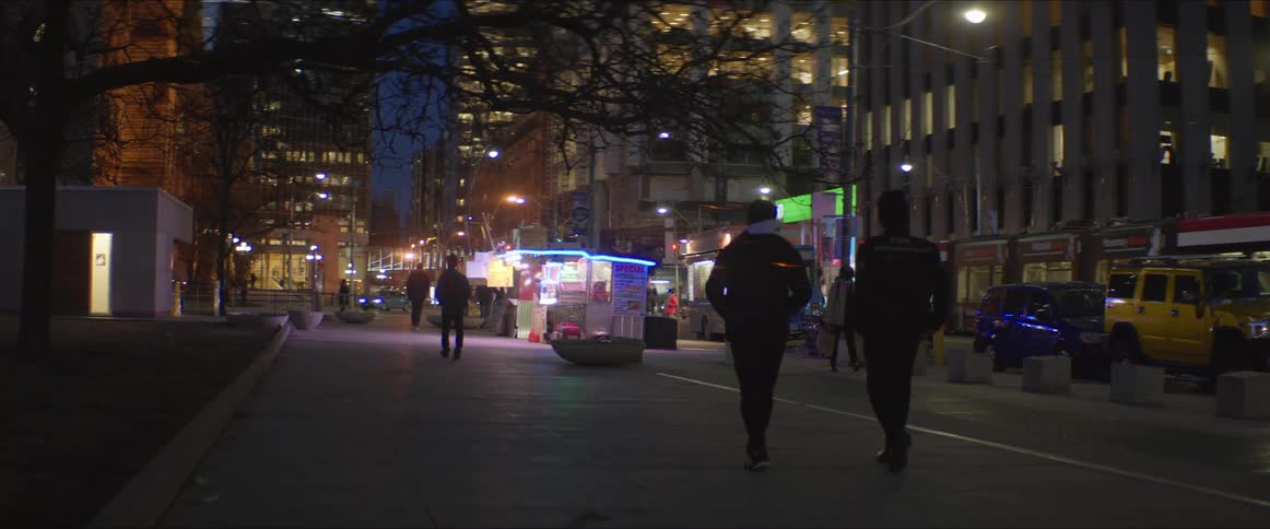 Pedestrians walking on a sidewalk in a city at nighttime next to a busy intersection surrounded by high rise buildings
