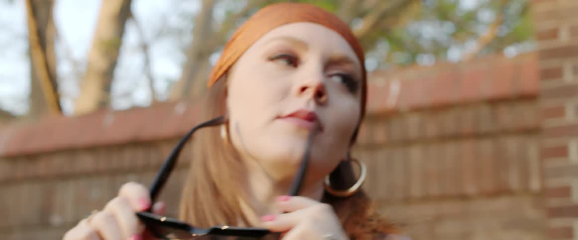 A woman removes sunglasses from her face and poses as she is photographed outdoors during the day