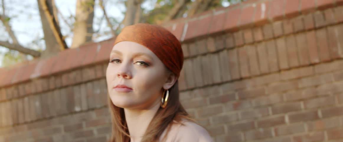 Woman wearing headscarf and earrings poses outdoors next to brick wall