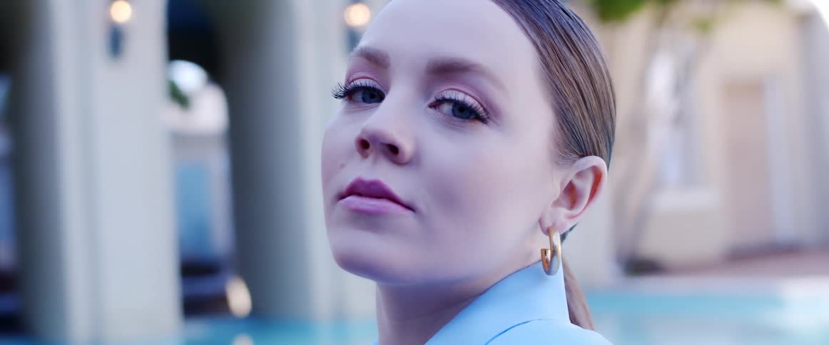 A woman in fashionable clothing and makeup gazes into the camera while standing outdoors during the day