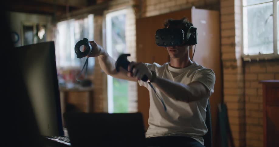 A man sits at a computer desk wearing a virtual headset playing with two vr remotes in his hand