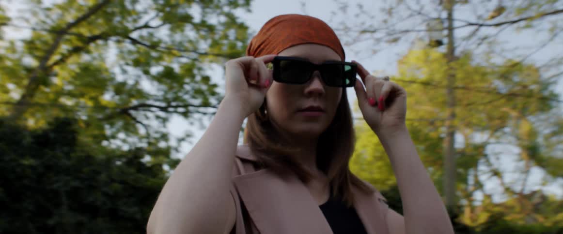 A woman with sunglasses getting pictures taken of her with trees the sky and clouds in the background