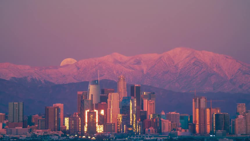 A city skyline with buildings with a mountain range the moon the sky and clouds in the background