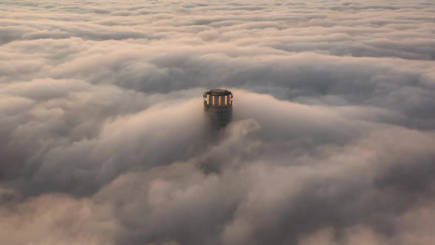 A building with windows surrounded by fog with the sky and clouds in the background