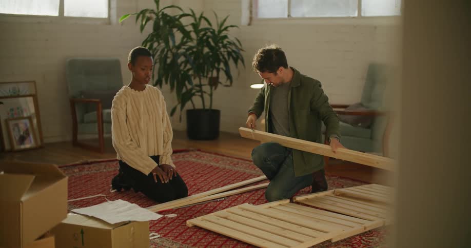 A woman speaks to a man as they assemble furniture while kneeling on a carpet in the living room of a house during the day