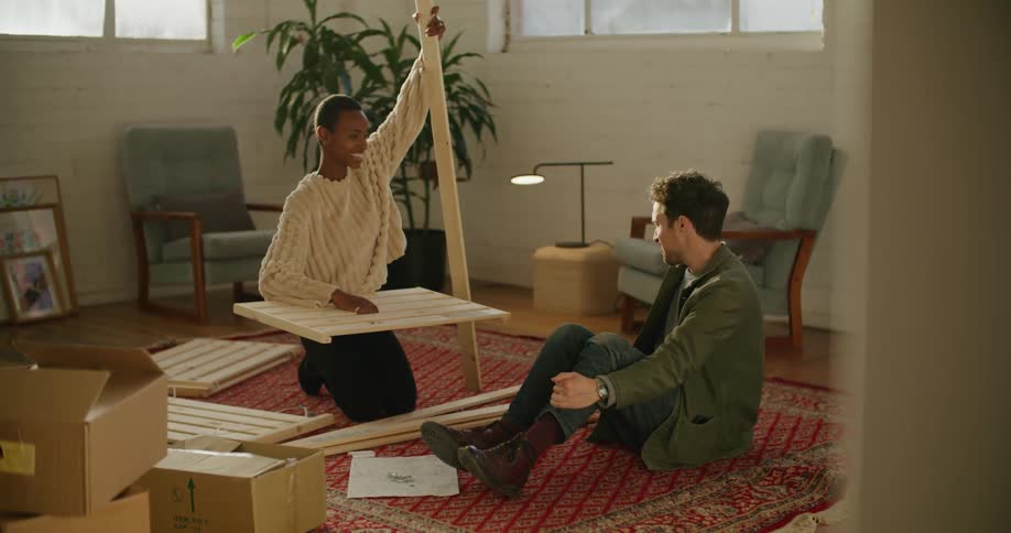 A woman laughs while assembling furniture alongside a man in the living room of a house during the day