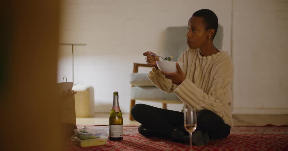 A young woman sitting on a rug eats from a bowl while listening to a person sitting across from her