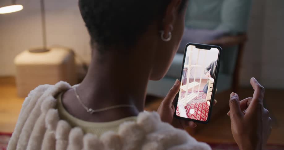 A woman standing looking at a phone screen instructing a man kneeling on a rug in a living room on how to put together a piece of furniture