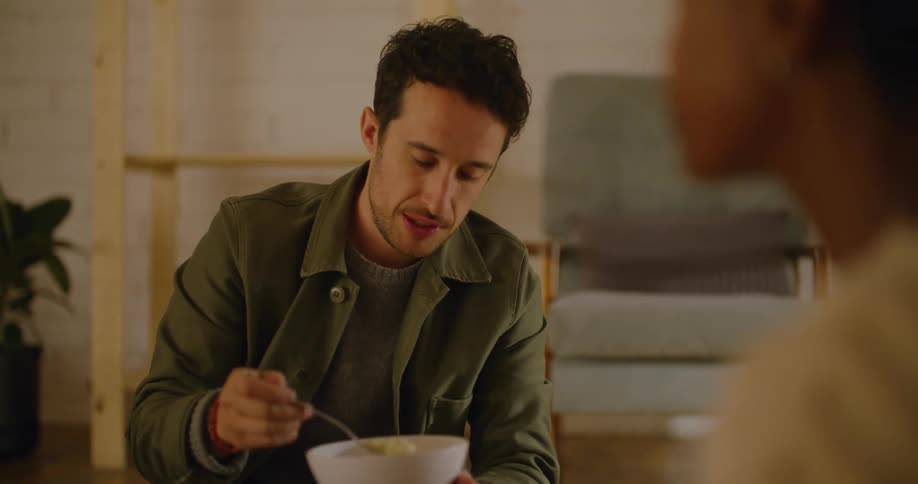 A man eats from a bowl chatting with a woman sitting on the floor across from him in the living room of a house