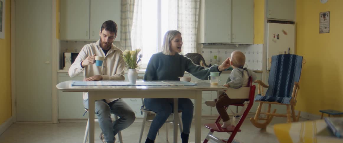 A man and woman sit at a kitchen table while feeding their baby seated in a highchair