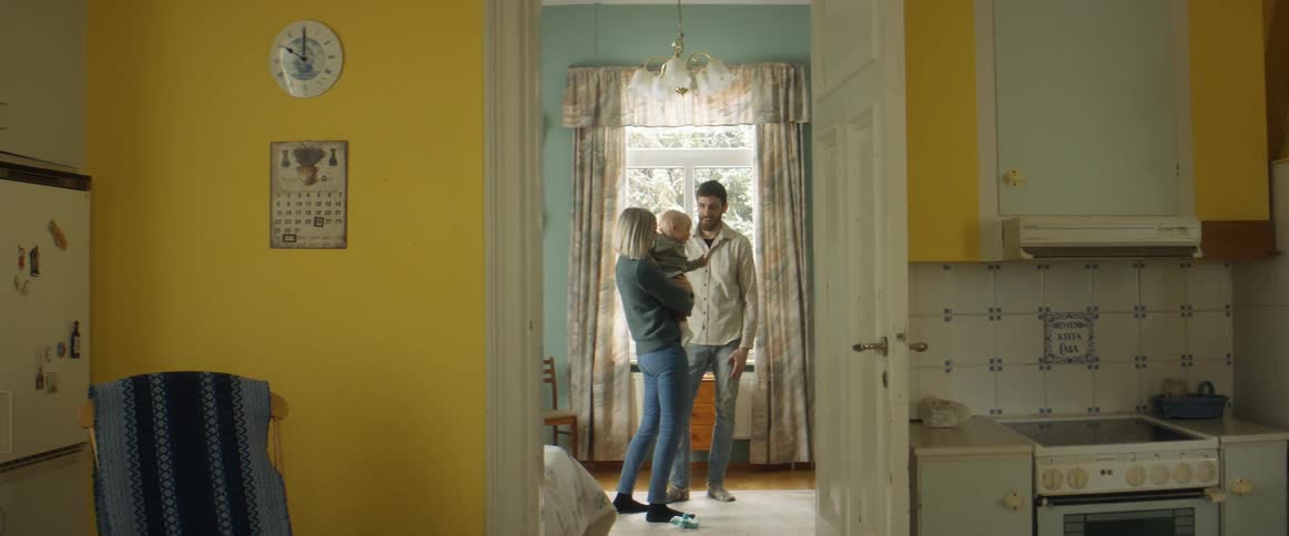 A man a woman and a baby in a room with a wall and window in the background