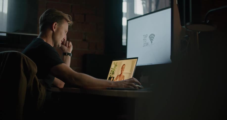 A man seated at a desk speaks to a woman via videoconferencing while he looks at a larger computer screen