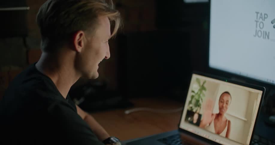 A man waves to a young woman through video chat on his laptop as he sits at a desk indoors