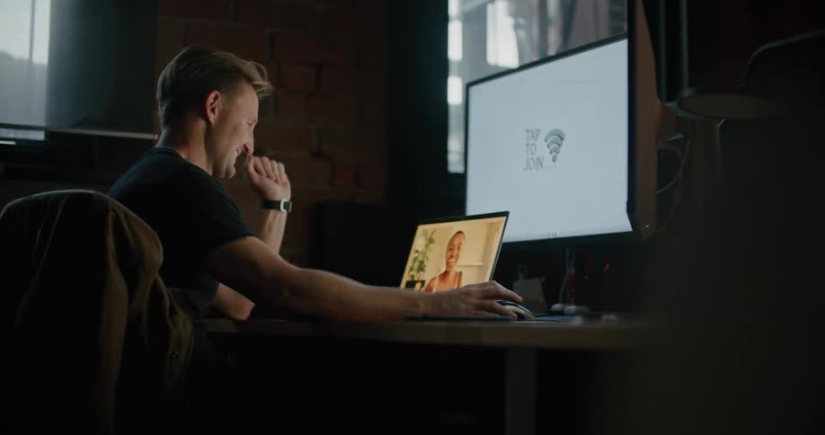 A man sits at his office at the office and waves at a colleague at home during a video call on his laptop