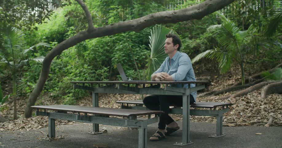 Man sitting outdoors at picnic table waves during video call on laptop