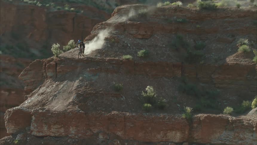 Person rides bicycle down narrow mountain ridge slope kicking up dust