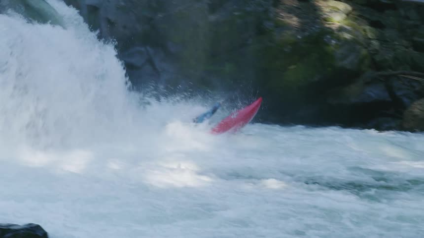 A man in a red whitewater kayak descends a waterfall in a dappled sun and moss covered river gorge