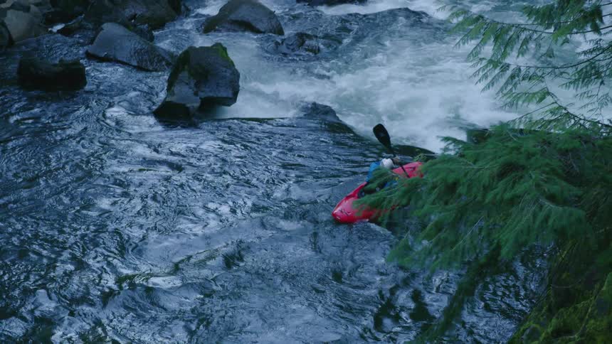 Real time shot from high above the whitewater below of a man in a red whitewater kayak paddling downstream away from camera at sundown