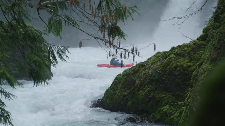 A man in a red whitewater kayak paddles in front of a huge waterfall in an impossibly scenic and clean river canyon