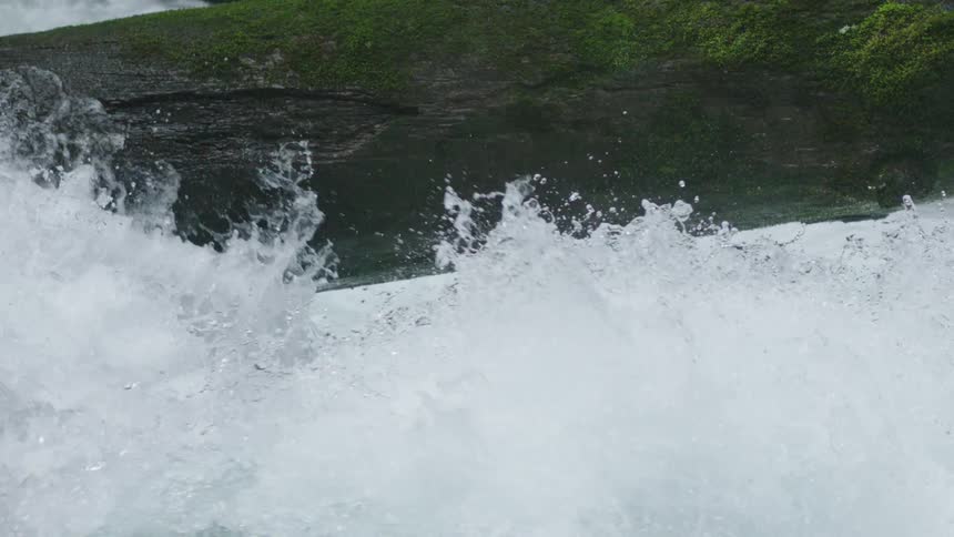 A view of rapids on a river in slow motion looking straight downstream as the water explodes outward towards a log over the river