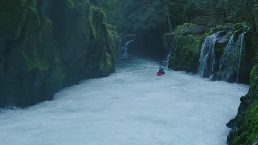 A man in a red whitewater kayak paddles downstream with incredibly scenic waterfalls splashing into the river below and mossy green canyon walls