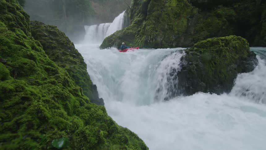 Handheld shot of a man paddling towards camera in a tight and dramatic river canyon descending waterfalls with cascading water all around him