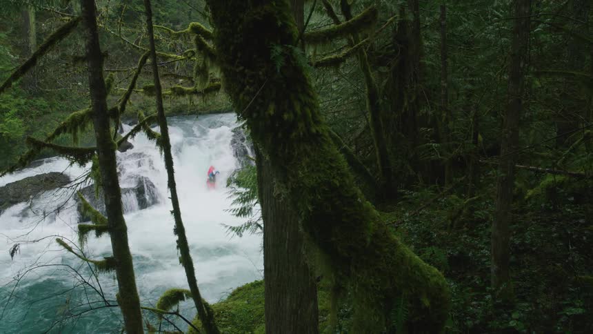 Superwide shot of a moss covered forest in the foreground and pristine clear whitewater in the background as a man in a red kayak descends a waterfall