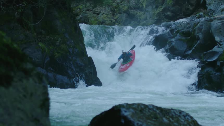 Slow motion shot of a man in a Red Whitewater Kayak paddling down a double waterfall and catching air in the middle and bottom of the drop