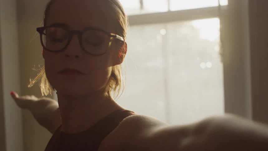 A woman stands in front of a window performing a yoga pose with her eyes closed and arms stretched