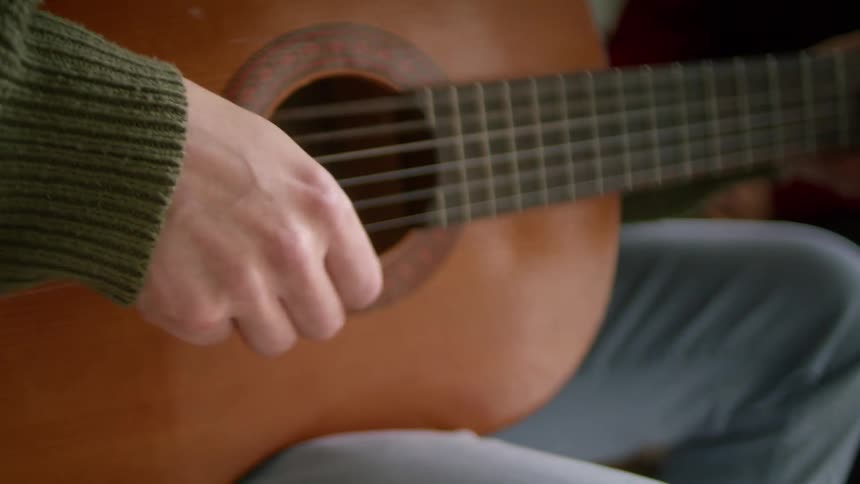 Woman in a red coat listens to man with a green sweater and grey jeans plays the guitar in the living room at home