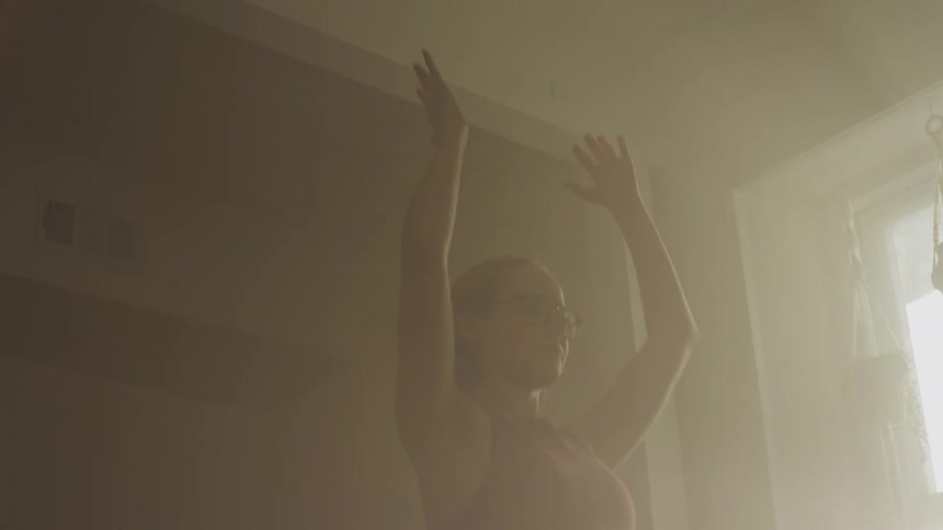 A woman standing indoors performing a yoga pose next to a window as daylight pours into the room