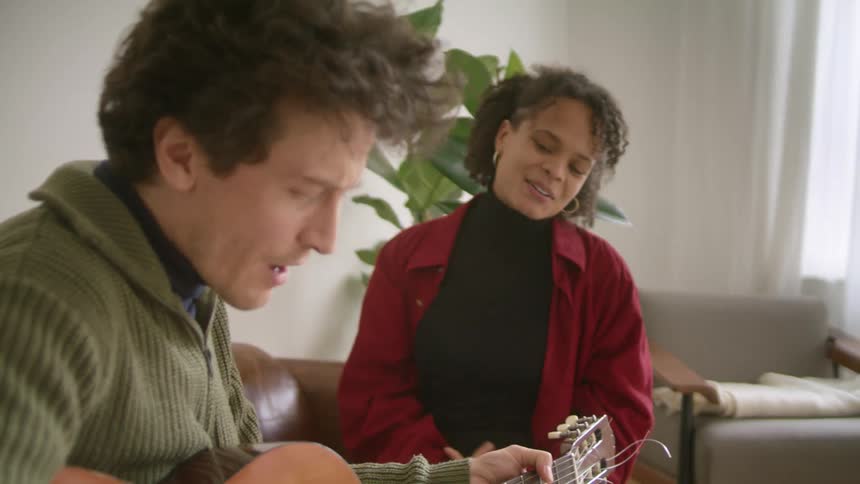 A man and woman singing and playing guitar on a living room sofa