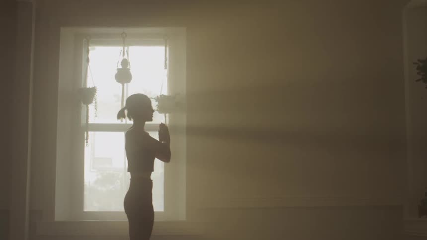 A silhouette of a woman stretching in a room with a wall and window in the background