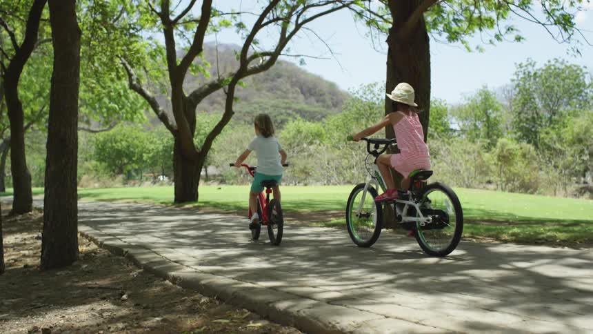 Two girls riding bikes on a pathway surrounded by trees with the sky and clouds in the background