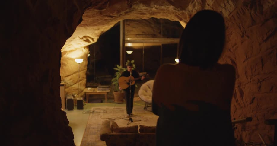 A woman stops in a cave corridor to wave a man play the guitar and sing in a living space created within a cave