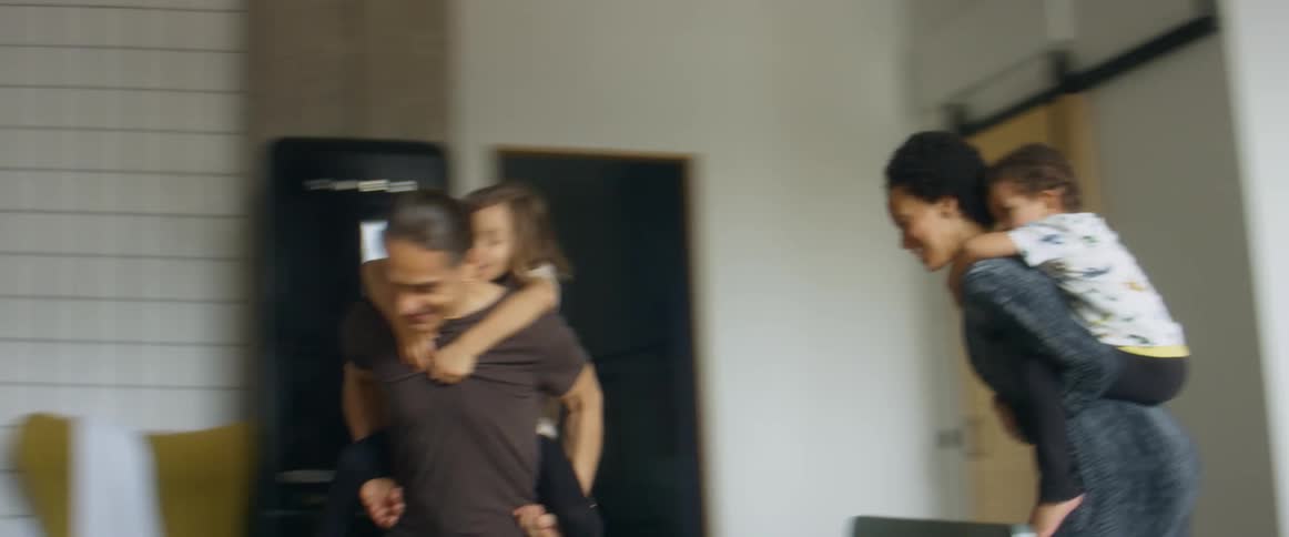 A man and woman with children on their back running around in a room with walls and windows in the background