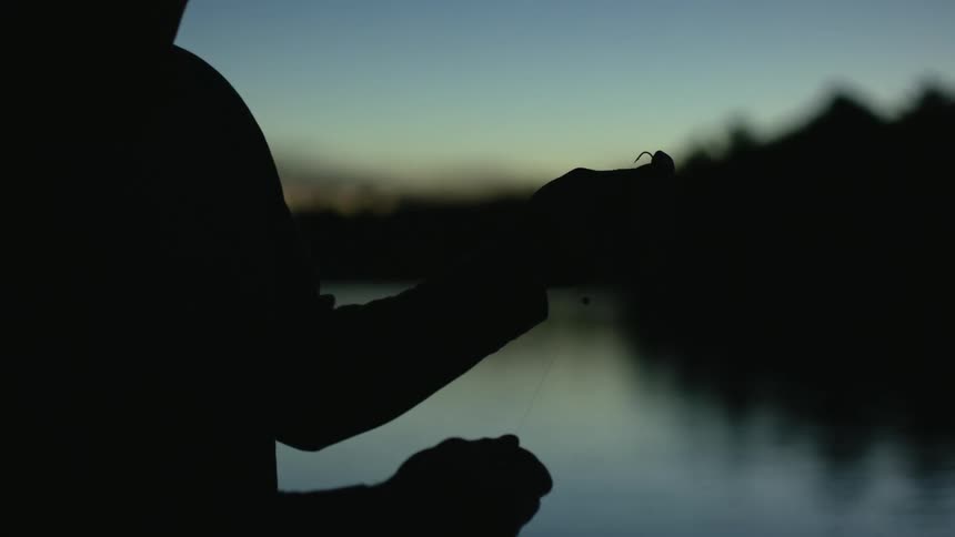 A silhouette of a person stretching out a fishing line with both hands next to a body of water in the evening