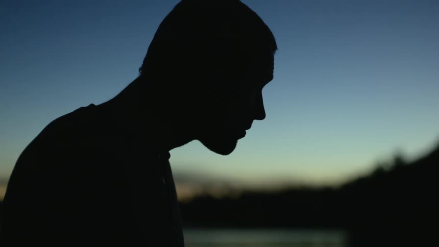 Silhouetted man standing outside raises bowed head