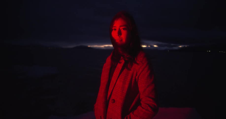Woman looking at the camera stands in a field at night lighted by a pulsing red light