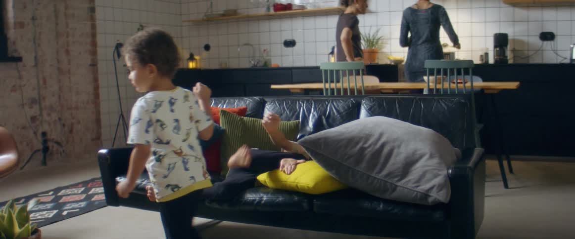 A brother and sister playfully fighting on a black couch in a living room while their parents cook in a kitchen at daytime