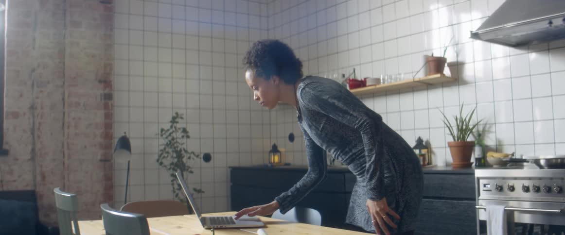 A woman cooking at home in her kitchen dances as she looks at her laptop and then goes back to her vegetables