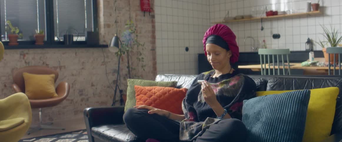 A black female wearing turban is sitting on the couch taking her temperature with a thermometer