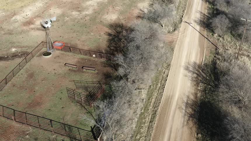 A dirt road passes by a small fence lined enclosure located on a dry and dusty farm